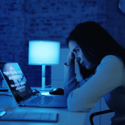 A dark blue photo of a woman looking upset at an open laptop.