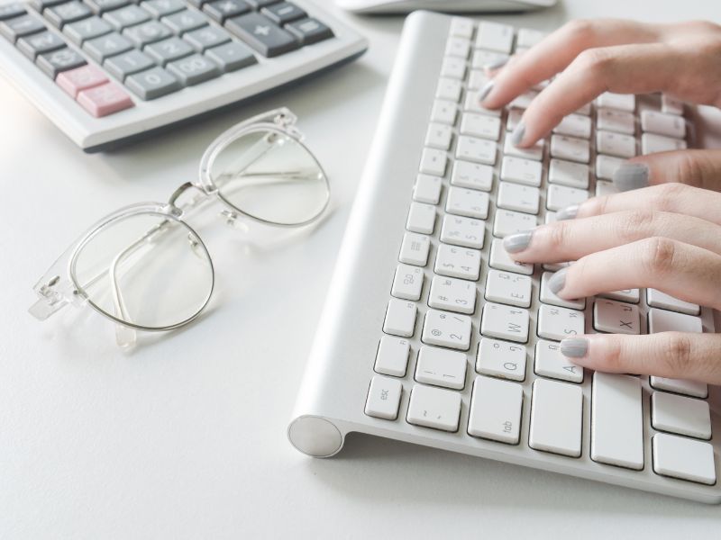 A close up of a white keyboard with hands about to type on the keys. A pair of reading glasses are nearby.